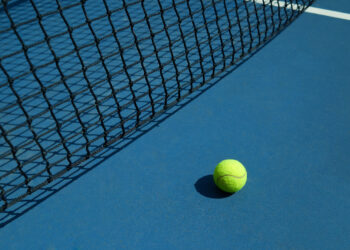 Yellow tennis ball is laying near black opened tennis court's net. Contrast image with satureted colors and shadows. Concept of tennis outfit photografing.