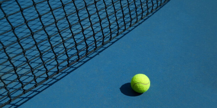 Yellow tennis ball is laying near black opened tennis court's net. Contrast image with satureted colors and shadows. Concept of tennis outfit photografing.